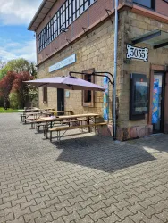 Eine Terrasse mit mehreren Tischen und Bänken vor einem Gebäude. Ein Sonnenschirm schützt vor der Sonne.