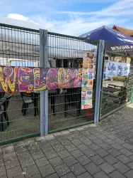 Ein einladendes Tor mit einem bunten "WELCOME"-Schild. Im Hintergrund sind Tische und Stühle sowie ein Sonnenschirm zu sehen.