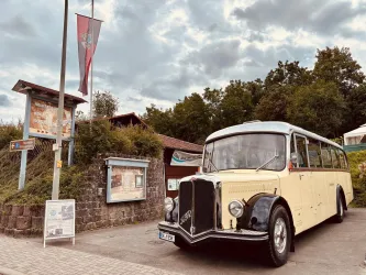 Ein nostalgischer Bus steht an einer Haltestelle, umgeben von Bäumen und einem grauen Himmel. Im Hintergrund sind Informationsschilder und eine Fahne sichtbar.