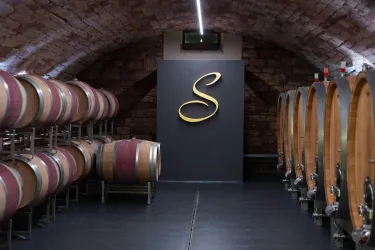 Ein Weinkeller mit großen Holzfässern und einer stilvollen Wand mit einem goldenen Logo. Die Atmosphäre ist rustikal und einladend.