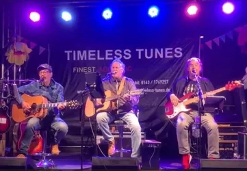 Ein Mann mit grauen Haaren steht mit einer Gitarre im Hintergrund. Er hat einen nachdenklichen Ausdruck und trägt ein einfaches schwarzes Oberteil.