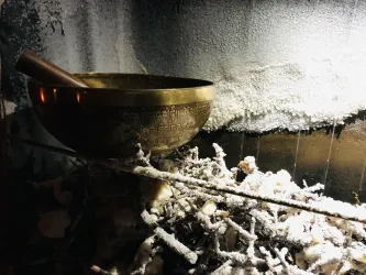 Eine Schale aus Metall steht auf einem Haufen von Ästen, umgeben von einer frostigen Umgebung. Der Hintergrund zeigt eine frostige Fläche, die dem Bild eine ruhige Atmosphäre verleiht.