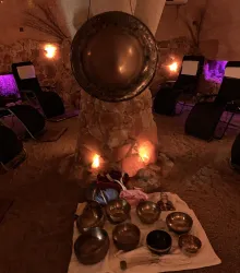 Ein entspannender Raum mit einer Sandfläche und leuchtenden Wänden. In der Mitte stehen verschiedene Klangschalen auf einem Tuch.