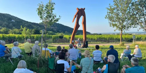 Eine Gruppe von Menschen sitzt auf Stühlen in einem Weinberg und schaut auf einen Musiker, der vor einer großen Skulptur spielt. Im Hintergrund sind sanfte Hügel und ein klarer Himmel zu sehen.