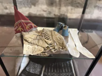 Auf dem Bild sind historische Textilien und Accessoires in einem Glasdisplay zu sehen. Dazu gehören Handschuhe, Socken und eine Kopfbedeckung mit bunten Mustern.