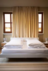 Ein komfortables Schlafzimmer mit einem großen, weißen Bett und sanften Vorhängen. Das Licht fällt durch die Fenster und schafft eine einladende Atmosphäre.