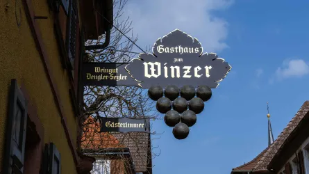 Ein Gasthaus mit dem Schild "Winzer" und Traubenmotiven. Im Hintergrund sind alte Gebäude und ein blauer Himmel sichtbar.