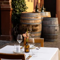 Ein schöner Restauranttisch mit Gläsern, Tellern und einer Vase mit Blumen. Im Hintergrund sind große Weinfässer zu sehen.