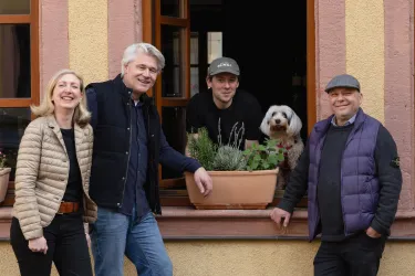 Eine Gruppe von vier Personen steht vor einem Fenster mit Pflanzkästen. Ein Hund schaut neugierig aus dem Fenster.