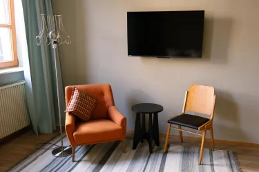 Ein gemütliches Wohnzimmer mit einem orangefarbenen Sessel und einem Holzstuhl. An der Wand hängt ein Fernseher, und der Boden ist mit einem gestreiften Teppich bedeckt.