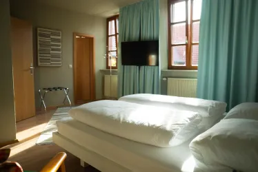 Ein modernes Schlafzimmer mit zwei Betten, hellen Farben und großen Fenstern. Ein Fernseher hängt an der Wand und sorgt für eine gemütliche Atmosphäre.