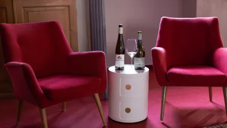 Zwei rote Sessel stehen neben einem runden Tisch. Auf dem Tisch stehen zwei Flaschen Wein und ein Weinglas.