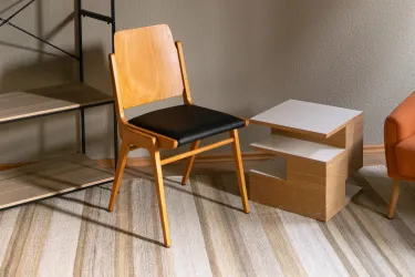 Ein moderner Holzstuhl mit schwarzem Sitzpolster steht neben einem kleinen Beistelltisch. Der Raum hat einen neutralen Farbton und einen gestreiften Teppich.