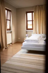 Ein heller und gemütlicher Schlafzimmerraum mit einem großen Bett und beigen Vorhängen. Der Holzboden und der Teppich verleihen dem Raum eine einladende Atmosphäre.