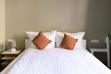 Ein modernes Schlafzimmer mit einem weichen Bett und zwei dekorativen Kissen. Neben dem Bett steht ein Nachttisch mit einer Lampe.