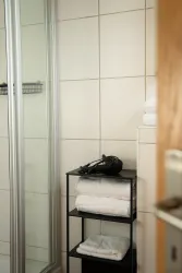 Ein modernes Badezimmer mit einer Glasdusche. Auf einem Regal liegen frische, gefaltete Handtücher und ein Haartrockner.