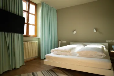 Ein schlichtes Schlafzimmer mit einem Doppelbett und hellen Vorhängen. Ein Fernseher ist an der Wand montiert und das Licht ist freundlich und einladend.