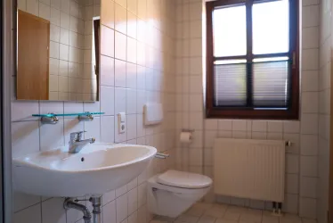 Ein einfaches Badezimmer mit einem Waschbecken, einer Toilette und einem Fenster. Die Wände sind mit weißen Fliesen gefliest.
