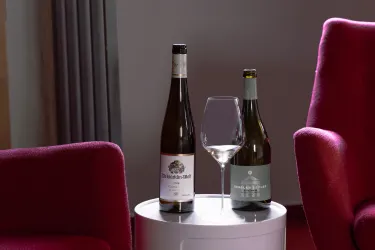 Auf einem Beistelltisch stehen zwei Weinflaschen und ein großes Weinglas. Die Flaschen sind unterschiedlich beschriftet und die Umgebung ist gemütlich mit roten Sesseln.