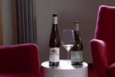 Zwei Weinflaschen stehen neben einem Weinglas auf einem Tisch. Die Umgebung ist modern mit roten Sesseln.