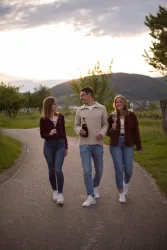 Drei junge Erwachsene spazieren entspannt auf einem Weg in der Natur. Sie halten jeweils eine Flasche in der Hand und genießen die Abendstimmung.