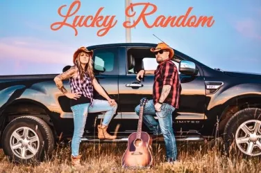 Ein Mann mit grauen Haaren steht mit einer Gitarre im Hintergrund. Er hat einen nachdenklichen Ausdruck und trägt ein einfaches schwarzes Oberteil.