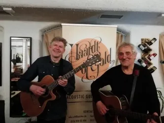Zwei Musiker stehen nebeneinander und halten akustische Gitarren. Im Hintergrund ist ein Banner mit dem Schriftzug "Lizard of Gold" zu sehen.