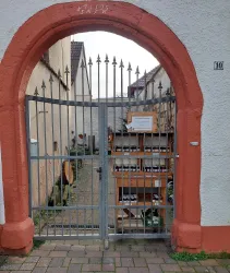 Ein schmiedeeiserner Torbogen mit einer Gittertür in einer schmalen Gasse. Hinter dem Tor befinden sich Holzregale mit Flaschen und Pflanzen.