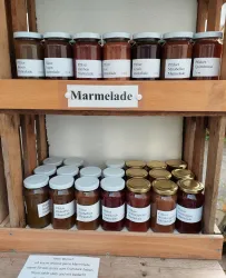 Ein Holzregal mit verschiedenen Marmeladengläsern in unterschiedlichen Farben. Die Gläser sind beschriftet und bieten eine Auswahl an Geschmäckern.