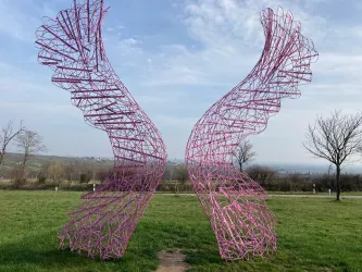 Eine kunstvolle Skulptur in Form von Flügeln steht in einer grünen Wiese. Der Himmel ist teilweise bewölkt und zeigt einen Hauch von Blau.