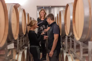 Drei Personen stehen in einem Weinkeller zwischen Eichenfässern. Sie halten Gläser und scheinen Wein zu probieren und zu diskutieren.