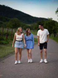 Drei junge Menschen spazieren lächelnd auf einem Weg. Die Umgebung ist grün und hügelig, ideal für einen entspannten Tag im Freien.