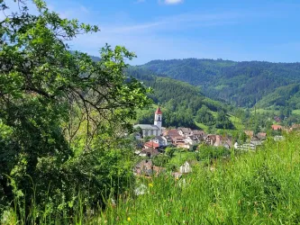 Ein malerisches Dorf in einer grünen Hügellandschaft. Im Hintergrund ist eine Kirche mit einem roten Turm sichtbar.