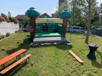 Eine farbenfrohe Hüpfburg in einem grünen Garten mit Bänken. Im Hintergrund sind Bäume und ein Gebäude sichtbar.