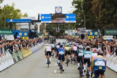 Eine Gruppe von Radfahrern fährt bei einem Wettkampf auf einer Straße. Viele Zuschauer stehen entlang der Strecke und verfolgen das Rennen.