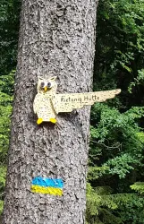 Ein Holzschild in Form einer Eule zeigt den Weg zur Rietania Hütte. Es ist an einem Baum befestigt, umgeben von grünen Blättern.