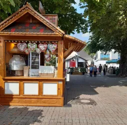 Ein Holzstand mit herzförmigen Lebkuchenherzen und einer Glasvitrine. Im Hintergrund sind Leute auf einem Platz zu sehen.