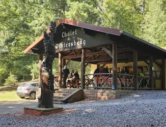 Ein Holzchalet im Wald mit der Aufschrift "Chalet du Wintersberg". Einige Besucher sind draußen versammelt, umgeben von grünen Bäumen.
