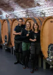 Drei Personen stehen lächelnd vor großen Holzfässern in einem Weinkeller. Alle halten ein Glas in der Hand und tragen schwarze Kleidung.