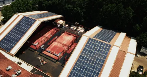 Zwei Gebäude mit Solarplatten auf dem Dach, die über Tennisplätze angeordnet sind. Der Bereich ist umgeben von Bäumen und hat eine moderne, nachhaltige Ausstrahlung.
