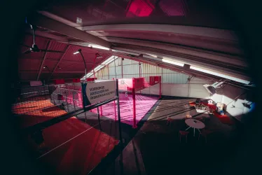 Eine moderne Sporthalle mit einem pinken Spielfeld. Es gibt Tische und Stühle für Zuschauer und eine freundliche Atmosphäre.