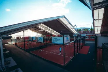 Ein Sportgelände mit mehreren Padel-Tennisplätzen unter einem modernen Dach. Die Courts sind umgeben von Anzeigen und es ist ein klarer Himmel zu sehen.