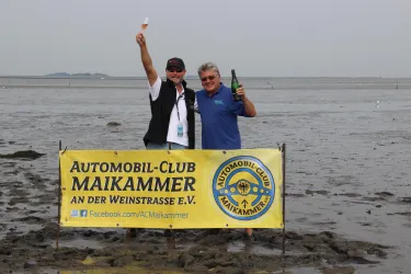 Zwei Männer stehen an einem Strand und feiern mit Getränken. Im Hintergrund ist ein Banner des Automobil-Clubs Maikammer sichtbar.