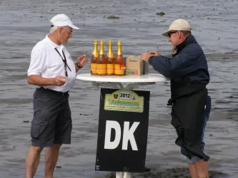 Zwei Männer stehen an einem Tisch im Wasser und unterhalten sich. Auf dem Tisch stehen mehrere Flaschen und ein Karton.