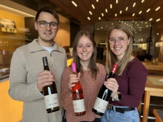 Drei Personen stehen in einem Weinlokal und halten Weinflaschen in der Hand. Sie lächeln und scheinen sich zu freuen.