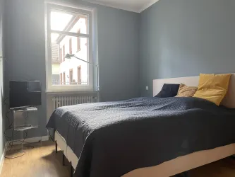 Ein gemütliches Schlafzimmer mit einem großen Boxspringbett und hochwertigen Desingelementen. Ein Fenster sorgt für Tageslicht und ein Fernseher steht ebenfalls bereit.