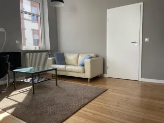 Eine Wohnecke mit einem hellen Sofa und einem Glastisch. Die Holzdielen verleihen. dem Raum eine gemütliche Atmosphäre, die einen spannenden Kontrast mit der modernen Einrichtung eingeht.