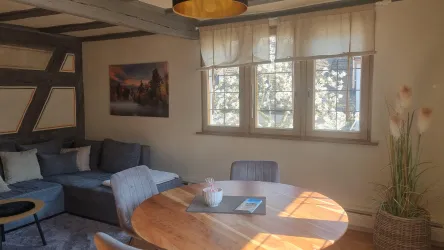 Ein gemütliches Wohnzimmer mit einem großen Sofa und einem runden Esstisch. Großzügige Fenster lassen viel Licht herein und zeigen die natürliche Umgebung.