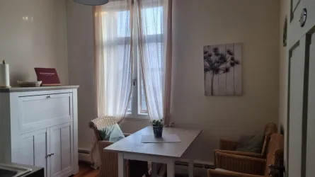 Ein heller Raum mit einem Tisch und drei Stühlen. Große Fenster lassen viel Licht herein und die Wände sind in neutralen Tönen gehalten.