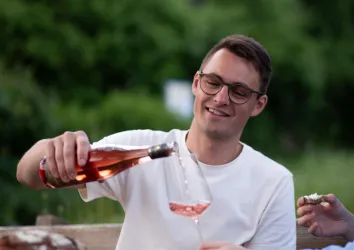 Ein Mann gießt Roséwein in ein Glas. Im Hintergrund sind grüne Bäume zu sehen.